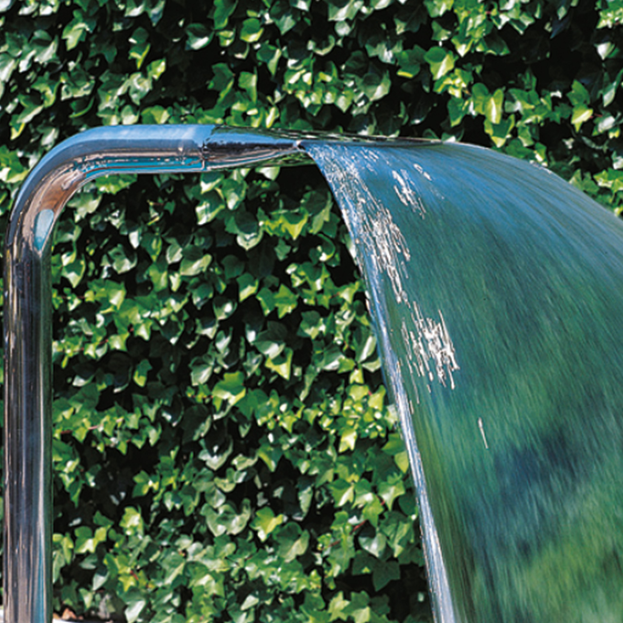 Breiter, gleichmäßiger Wasservorhang des Schwallkörpers Boa V4A aus glänzendem Edelstahl vor grüner Hecke.