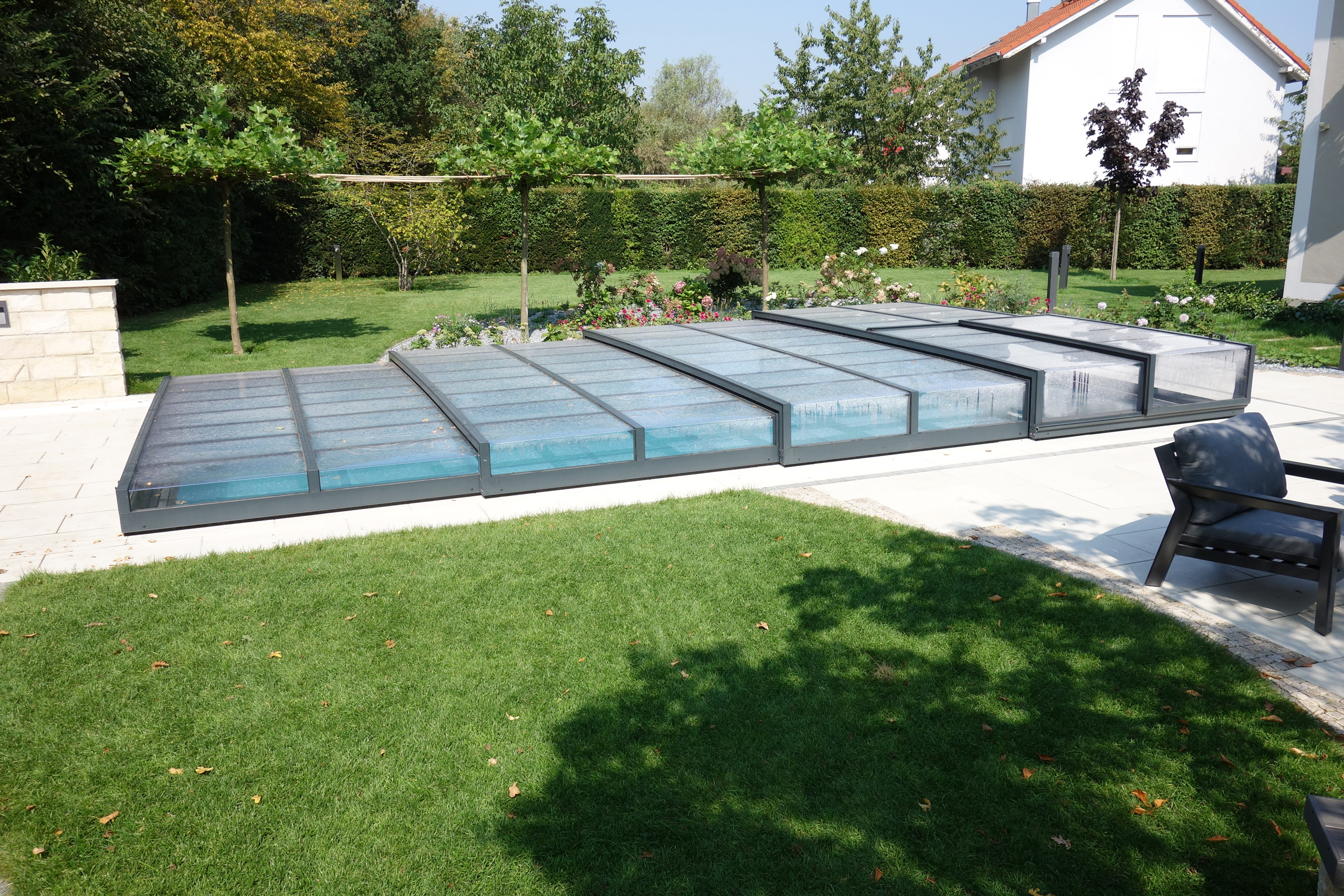 Überlaufbecken Overflow Stone im Garten, umgeben von Rasen und Pflanzen, mit einer Poolüberdachung.