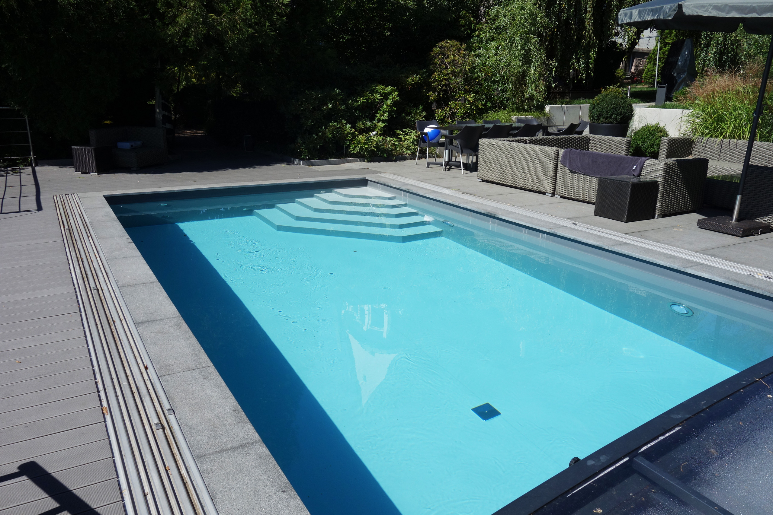 Betonbecken mit Ecktreppe und Sitzbank, umgeben von einer Terrasse und Gartenmöbeln.