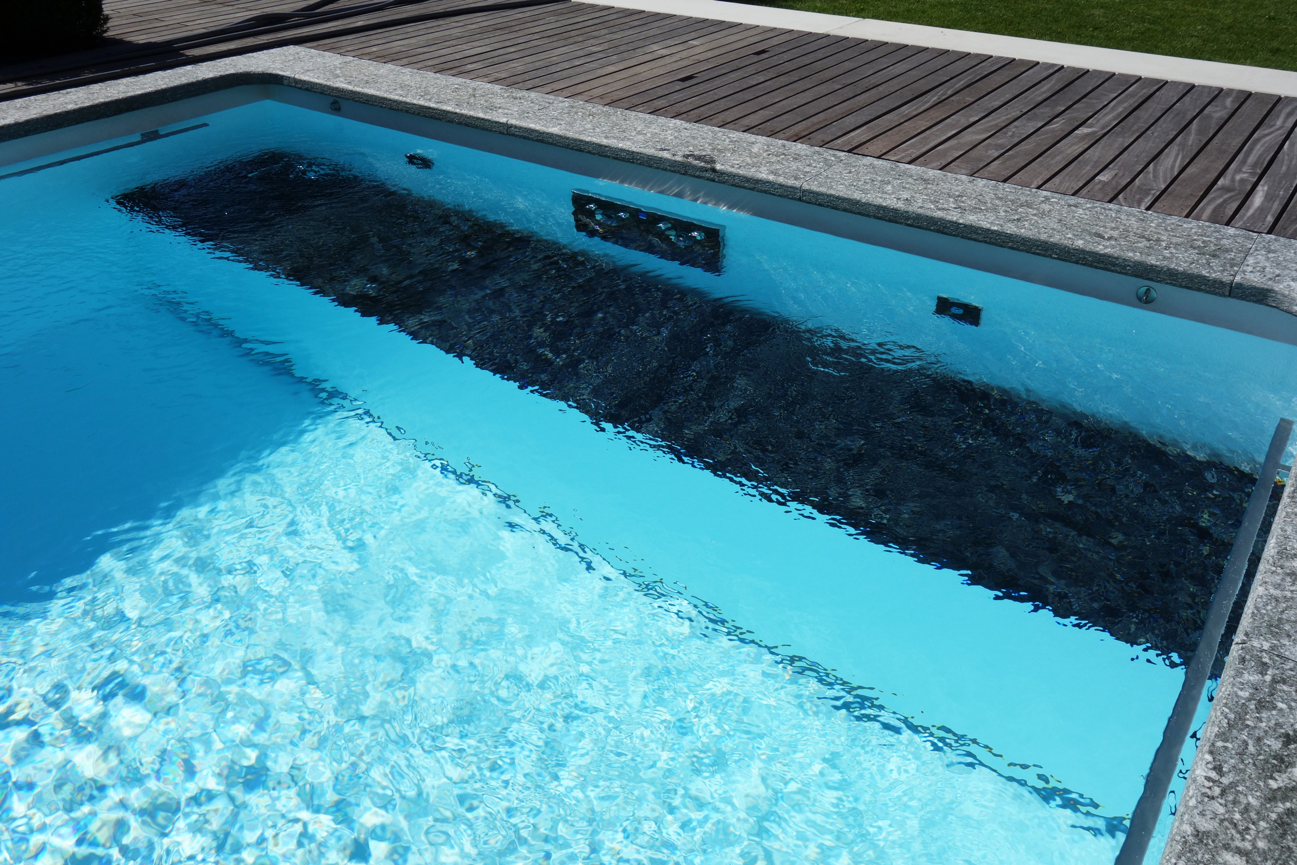 Blick auf die Unterflurabdeckung eines Rechteckbeckens mit Ecktreppe und Podest, umgeben von Holz und Stein.