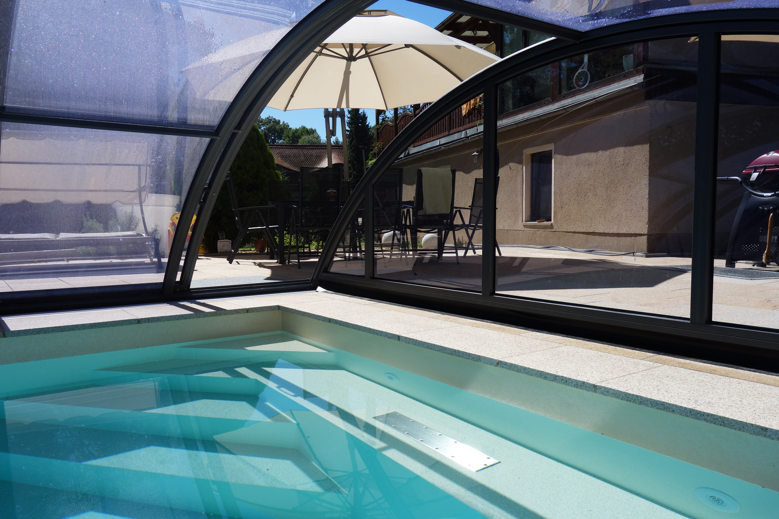 Blick aus dem Pool auf die Überdachung Klasik Clear und die Terrasse mit Sonnenschirm