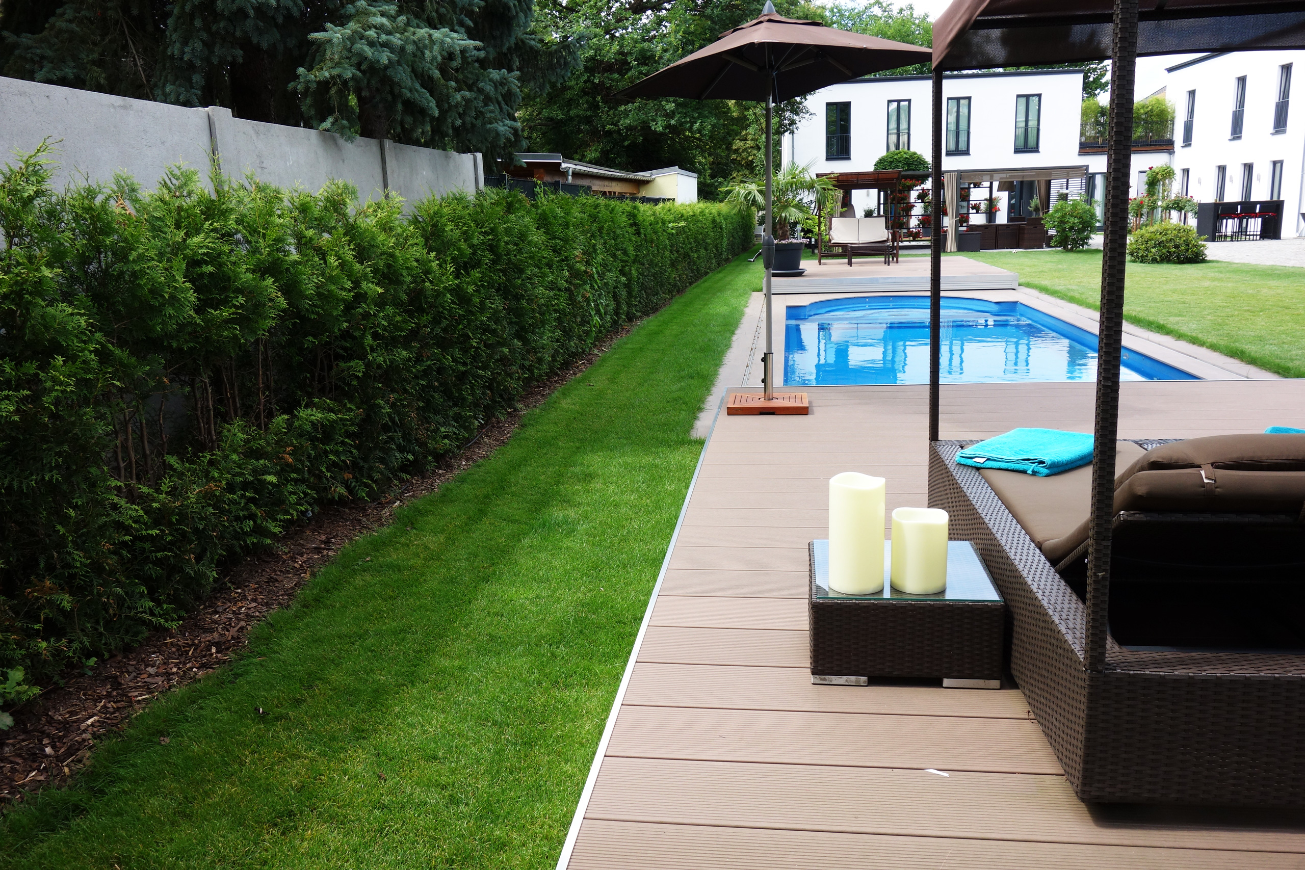 Braune Terrassendielen mit Pool, Liege und Kerzen, flankiert von einer Hecke und Rasen