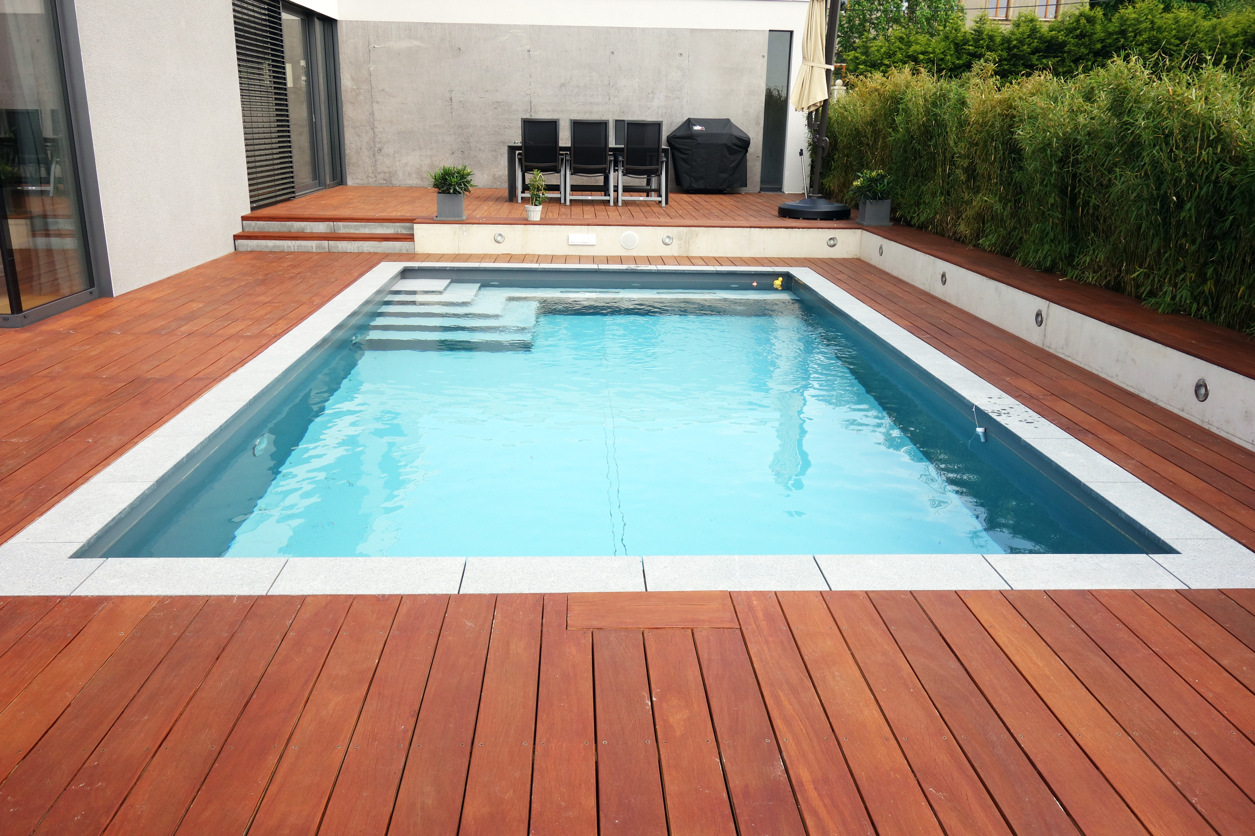 Betonbecken 3,5x7x1,5m mit Ecktreppe und Podest, umgeben von Holzterrasse und Bepflanzung
