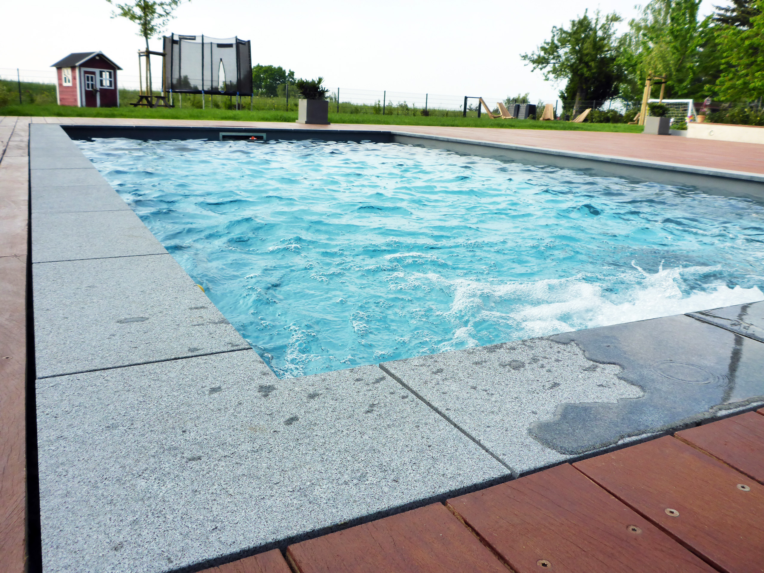 Sprudelndes Wasser im Betonbecken mit Ecktreppe und Podest, umgeben von grauen Platten und Holzterrasse