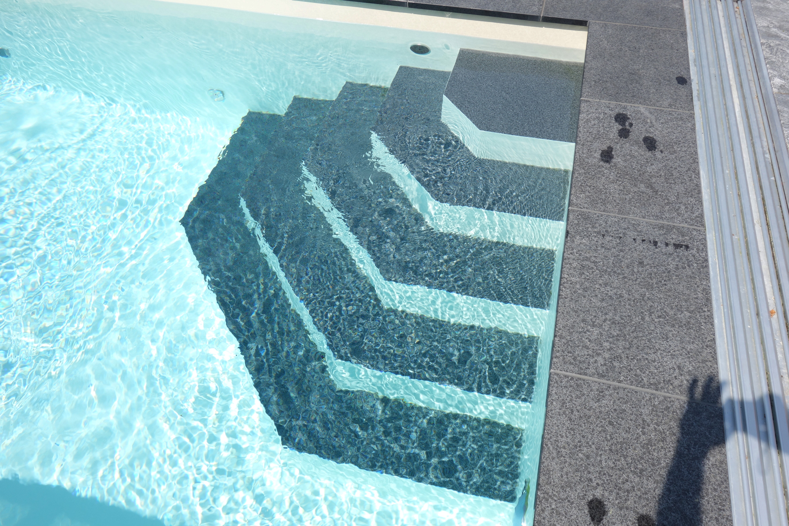 Ecktreppe im Außenbecken mit blauem Wasser, umgeben von dunklen Fliesen und einer Poolüberdachung