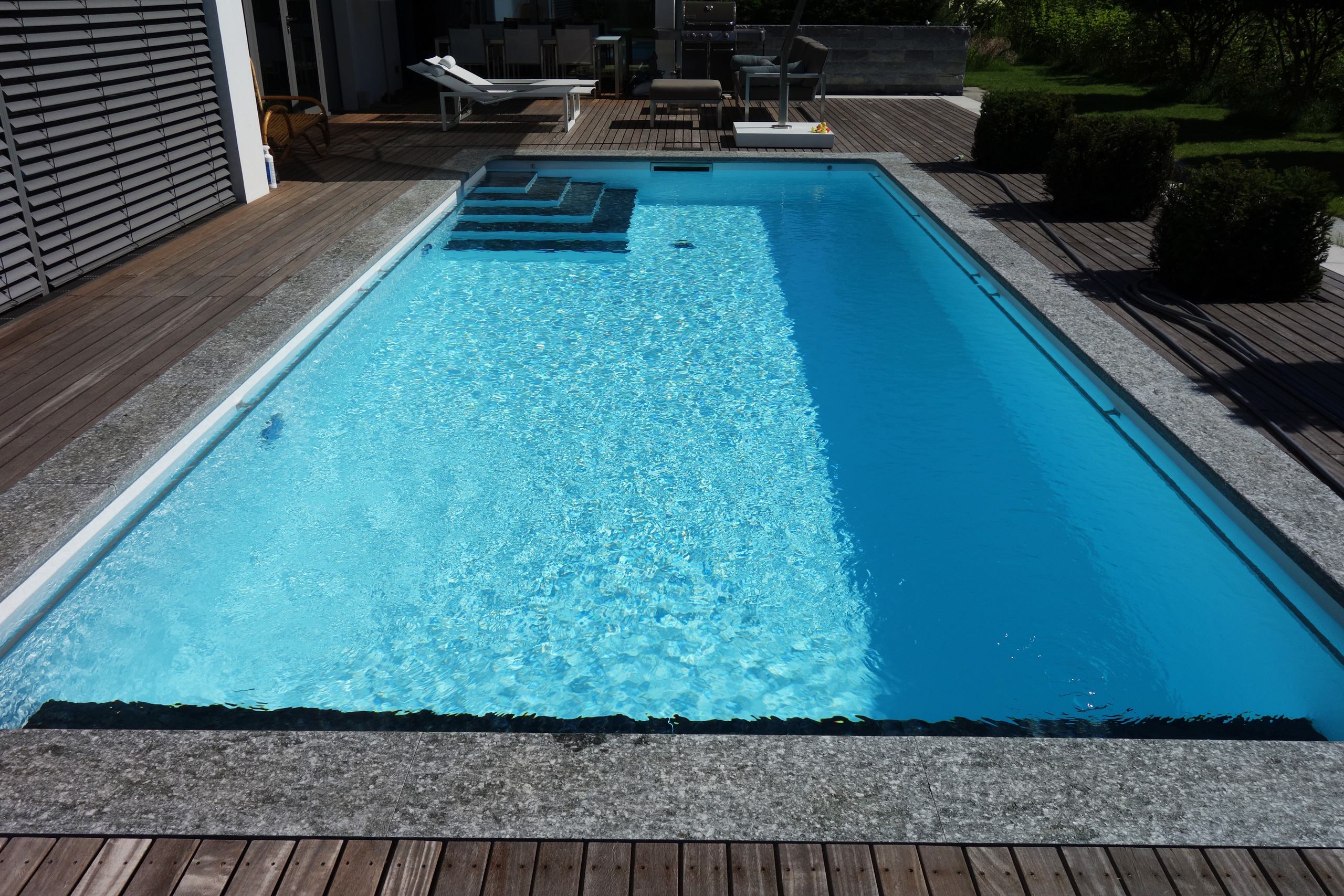 Rechteckbecken mit blauer Folie, Ecktreppe und Unterflurabdeckung, umgeben von Holzdeck und Steinrand