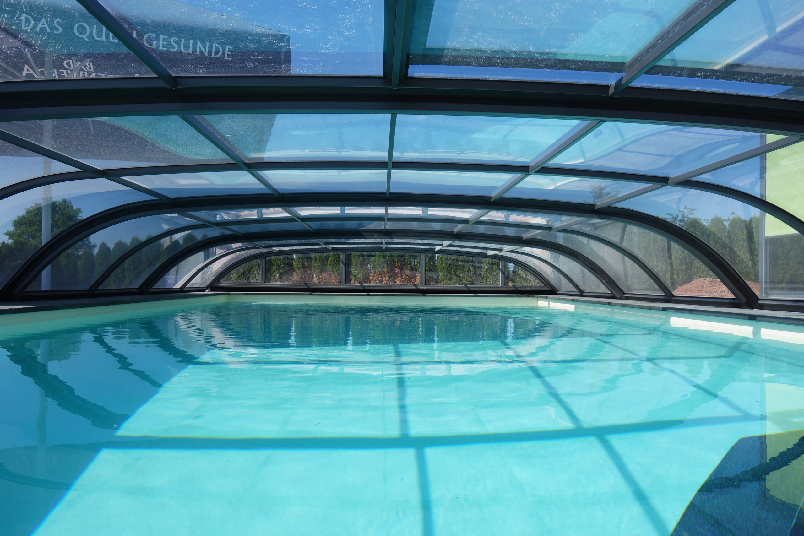 Blick in ein Rechteck-Schwimmbad mit hellblauer Folie unter einer transparenten Poolüberdachung.