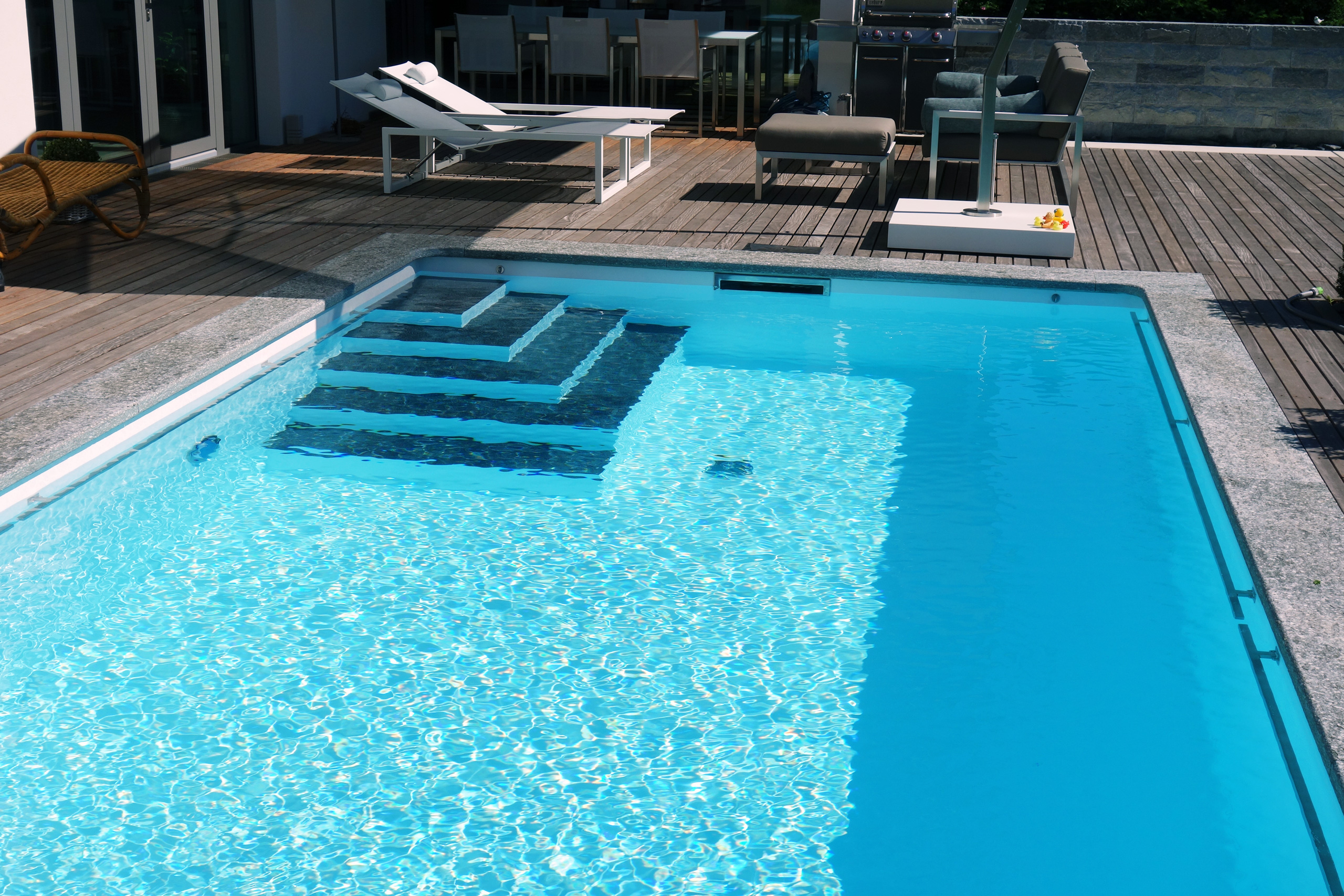 Blick auf das Rechteckbecken mit Ecktreppe, Podest und Unterflurabdeckung, umgeben von einer Holzterrasse.