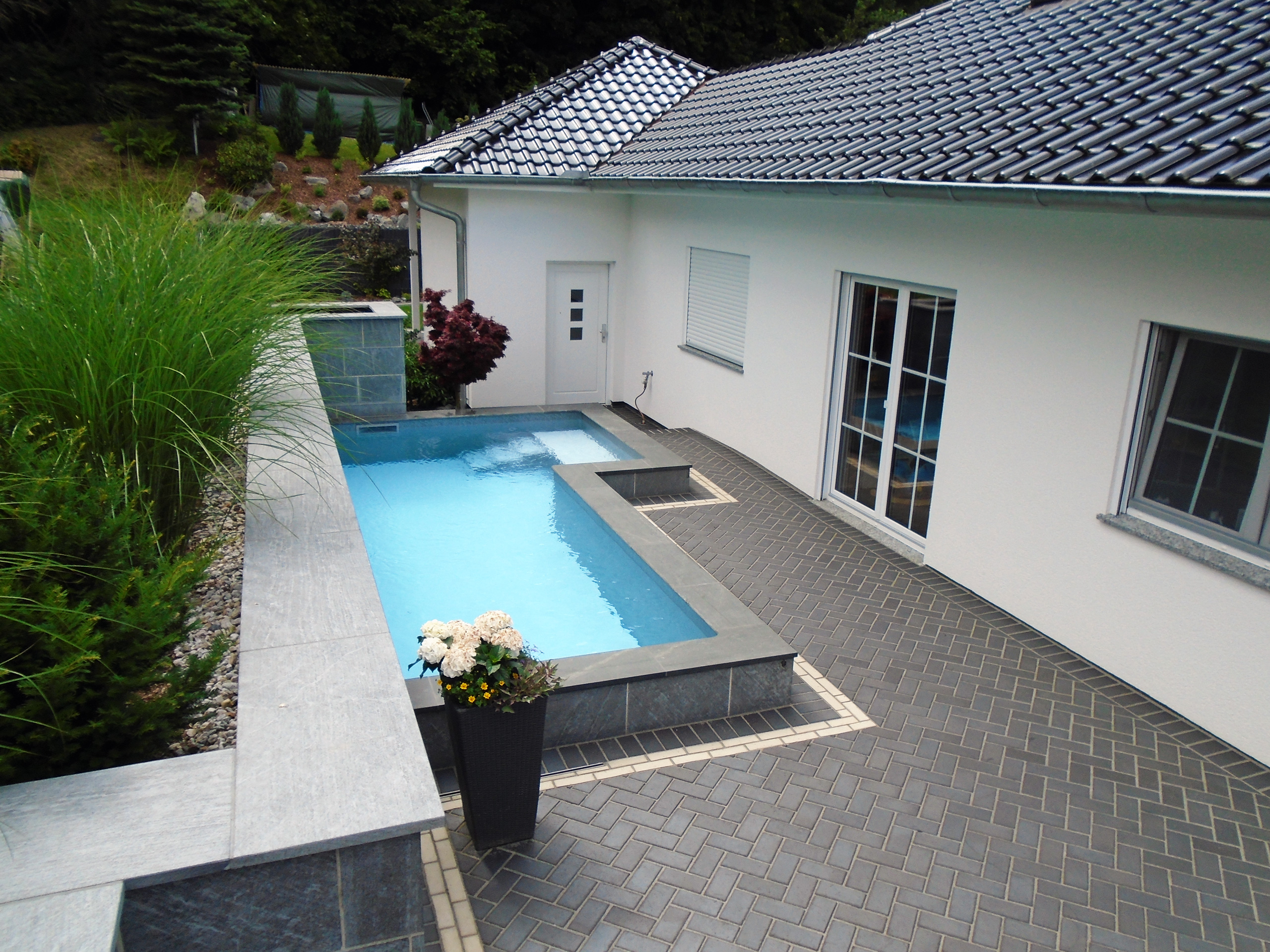 L-förmiges Schwimmbecken mit Treppe und Podest, umgeben von grauen Fliesen und Pflanzen.