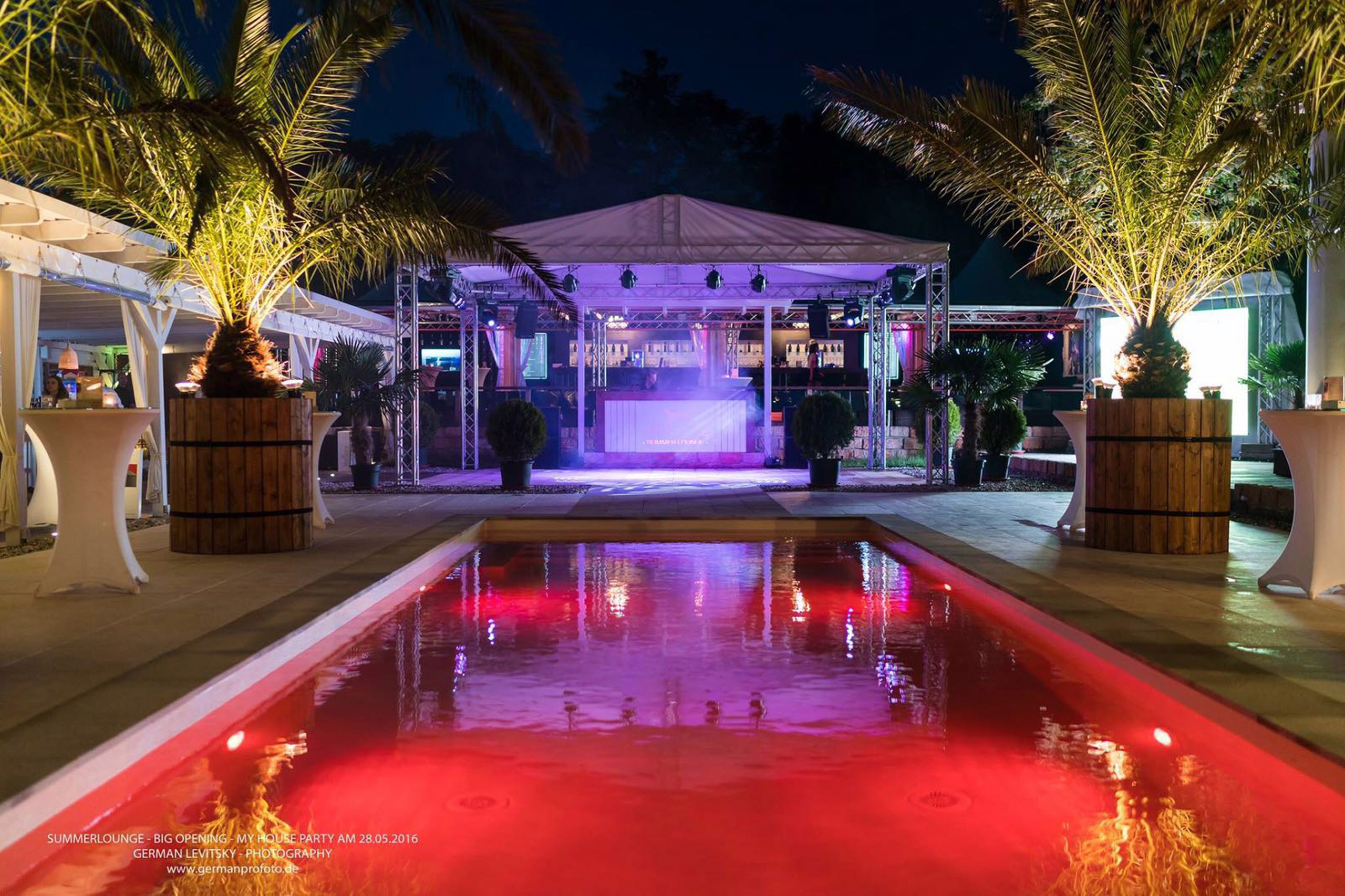 Längliches Schwimmbad mit roter Unterwasserbeleuchtung bei Nacht, umgeben von Palmen und einer Bar.