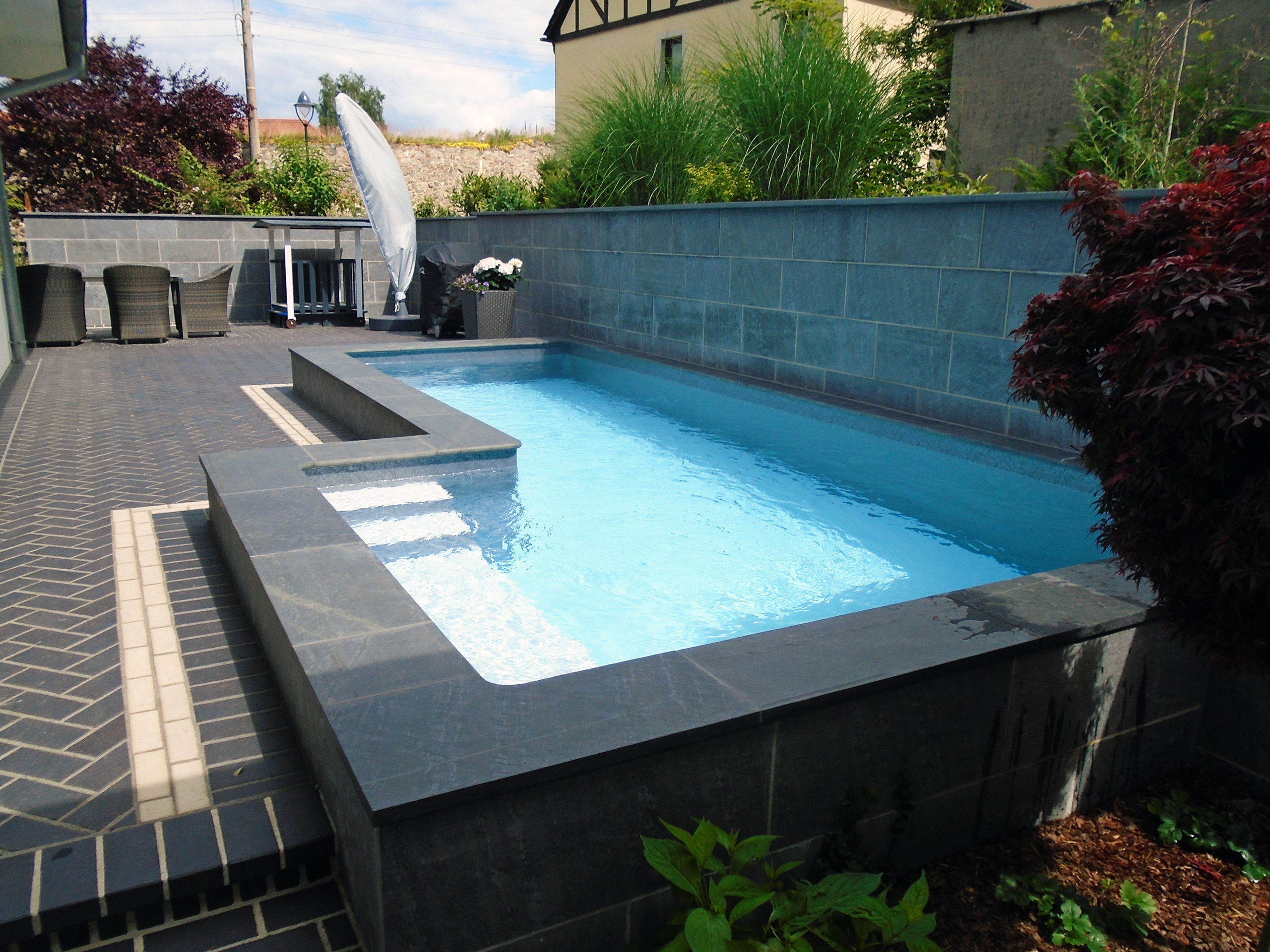 L-förmiges Schwimmbecken mit integrierter Treppe und Podest, umgeben von dunklen Fliesen und Pflanzen.