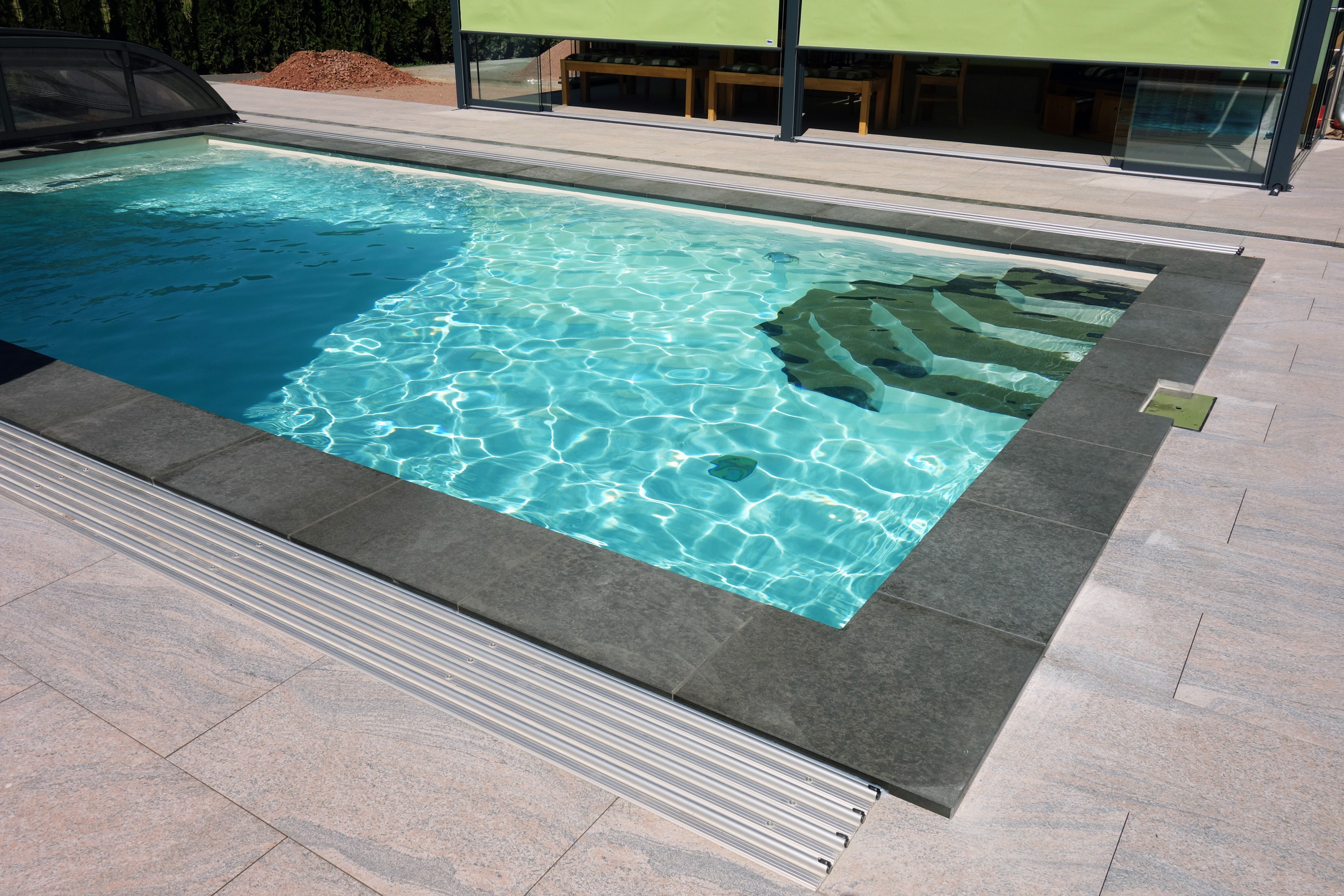 Rechteck-Schwimmbad mit Treppe, Gegenstromanlage und Abdeckung, umgeben von dunklen und hellen Fliesen