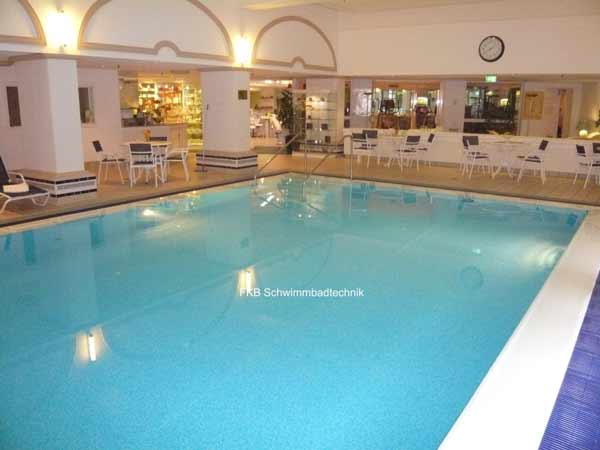 Großes Schwimmbecken mit Überlaufrinne in einer Hotelhalle, umgeben von Sitzgelegenheiten und Säulen.