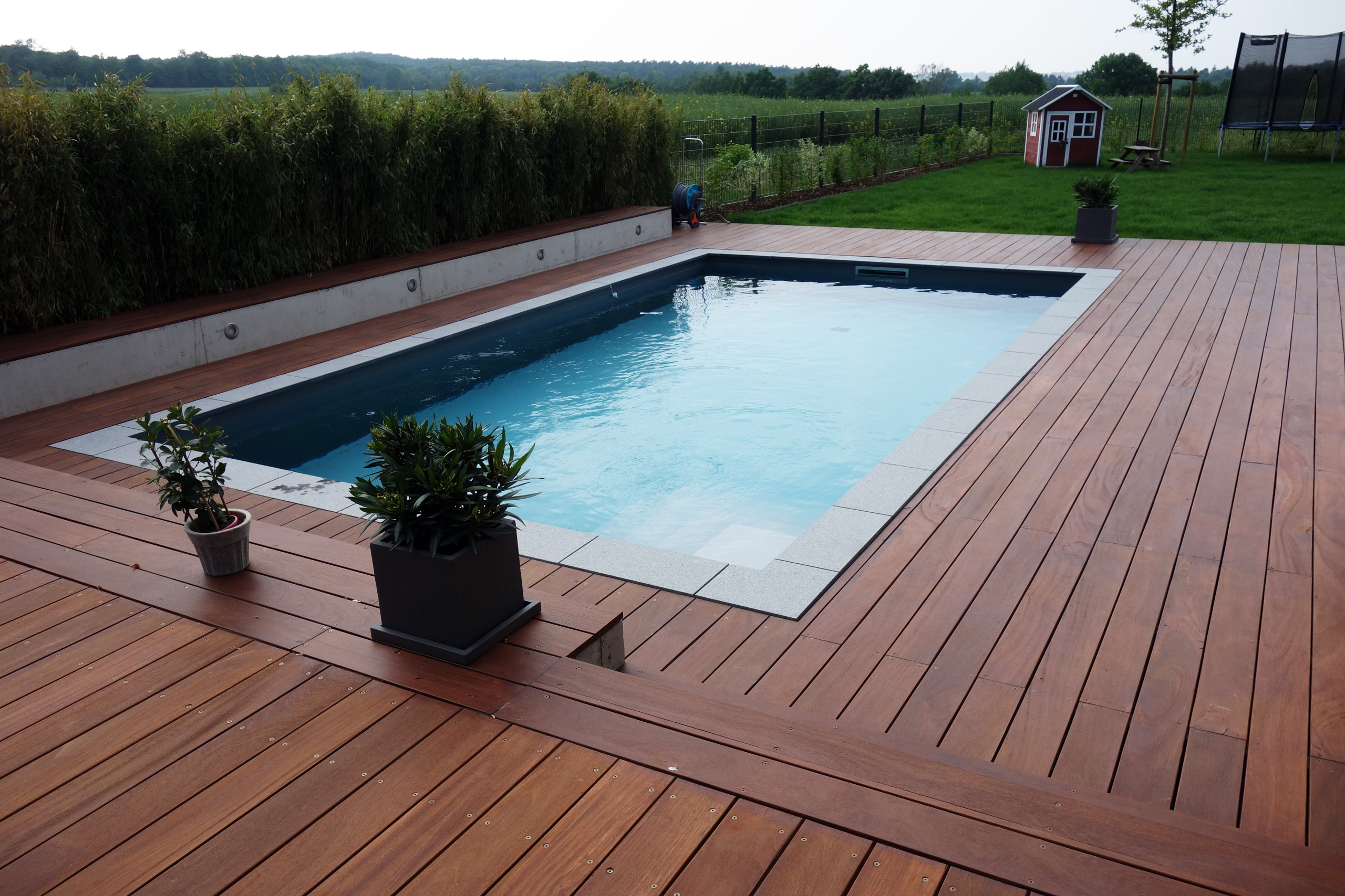 Betonbecken 3,5x7x1,5m mit Ecktreppe und Podest, umgeben von Holzterrasse und Bepflanzung