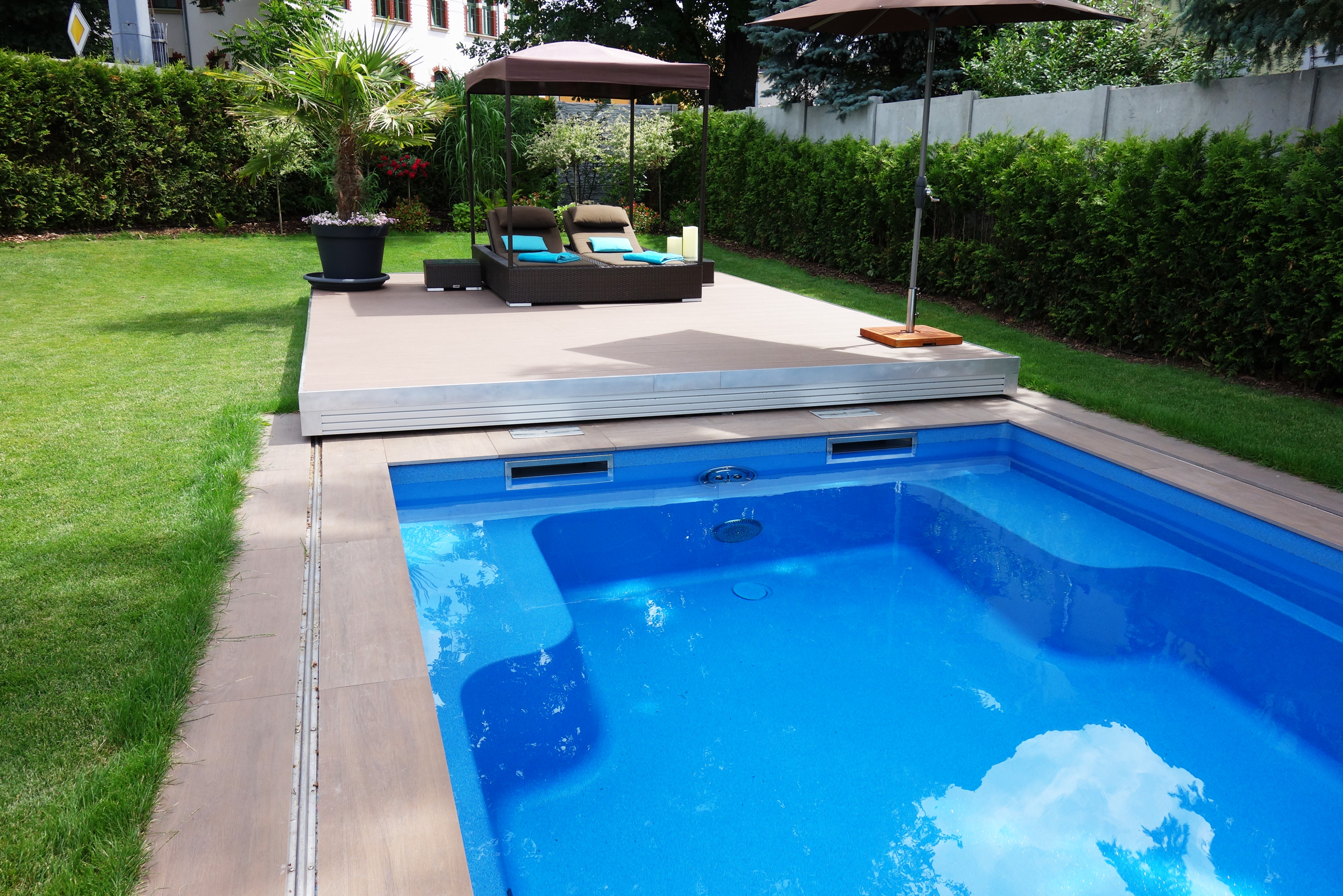 Blaues Compass Pool Schwimmbecken Java 101 mit geöffneter Terrassenabdeckung und Liegefläche im Garten