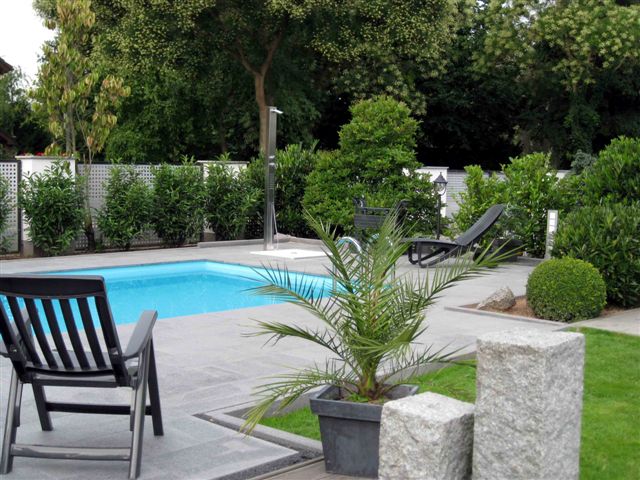 Garten mit türkisem Schwimmbecken, umgeben von grauen Steinplatten, Pflanzen und Liegen.