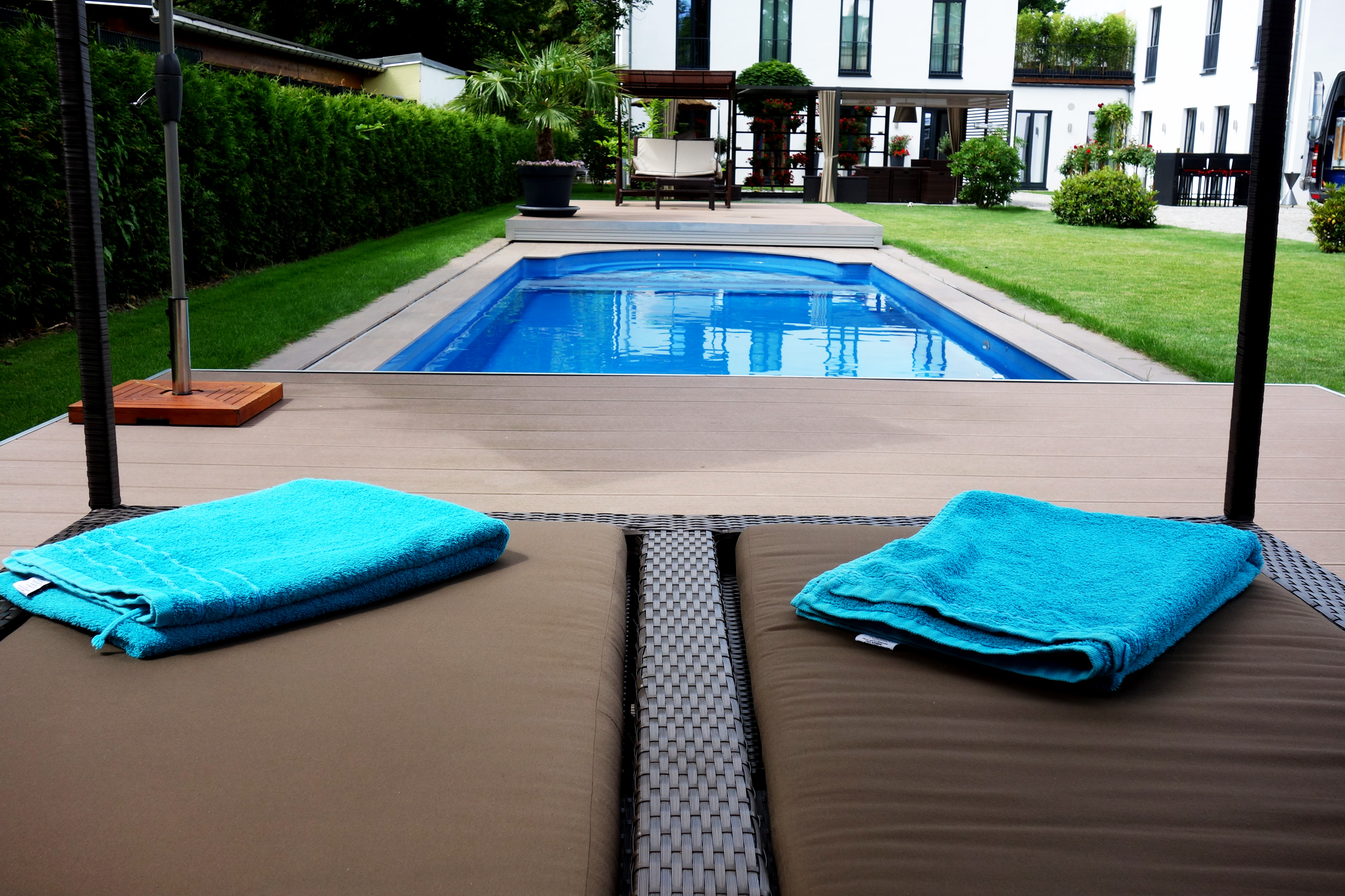 Blick über zwei Liegestühle mit blauen Handtüchern auf ein Compass Pool Schwimmbecken mit Terrassenabdeckung
