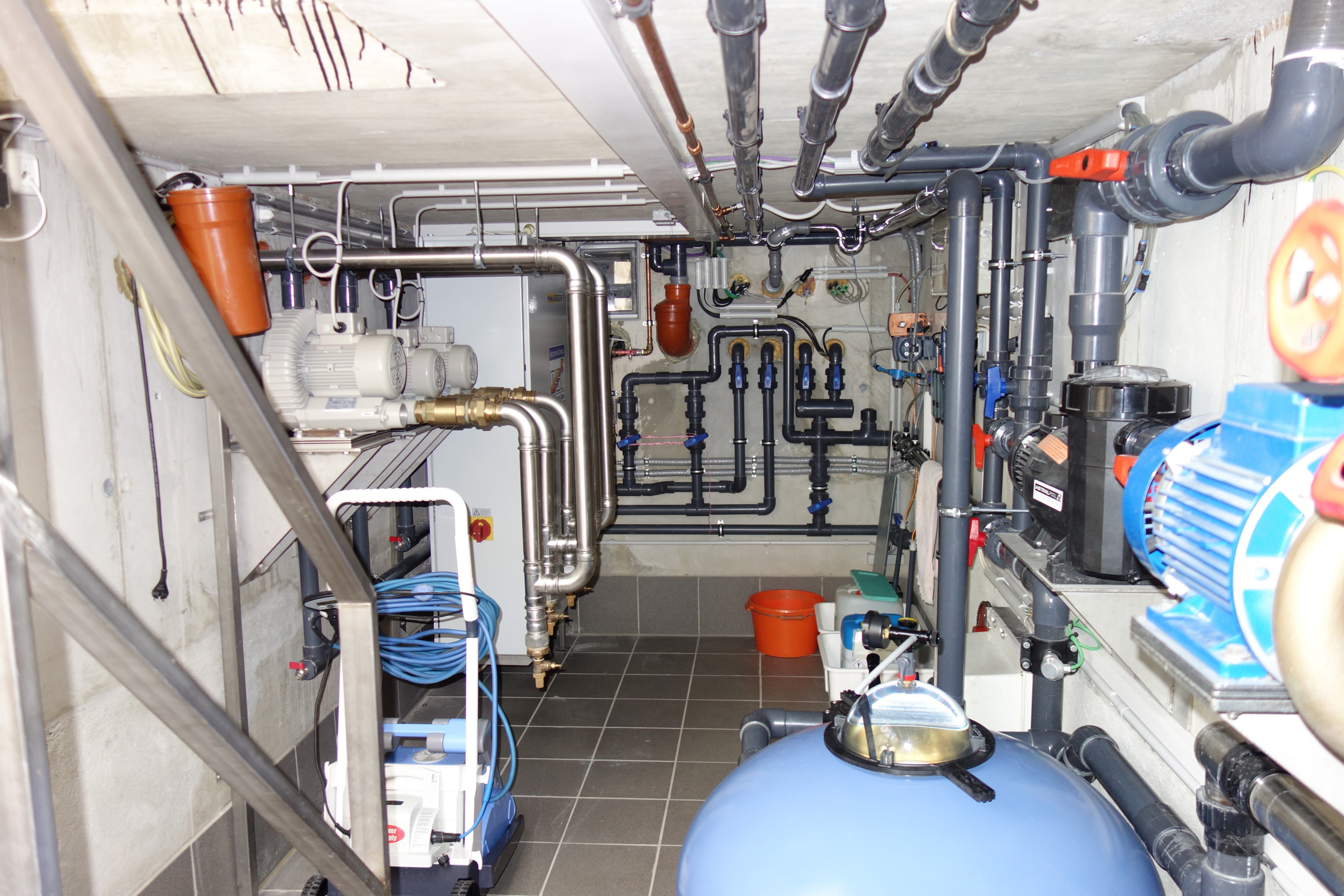 Blick in einen Technikraum mit blauen Filterkesseln, Pumpen, Rohren und Ventilen für ein Schwimmbecken.