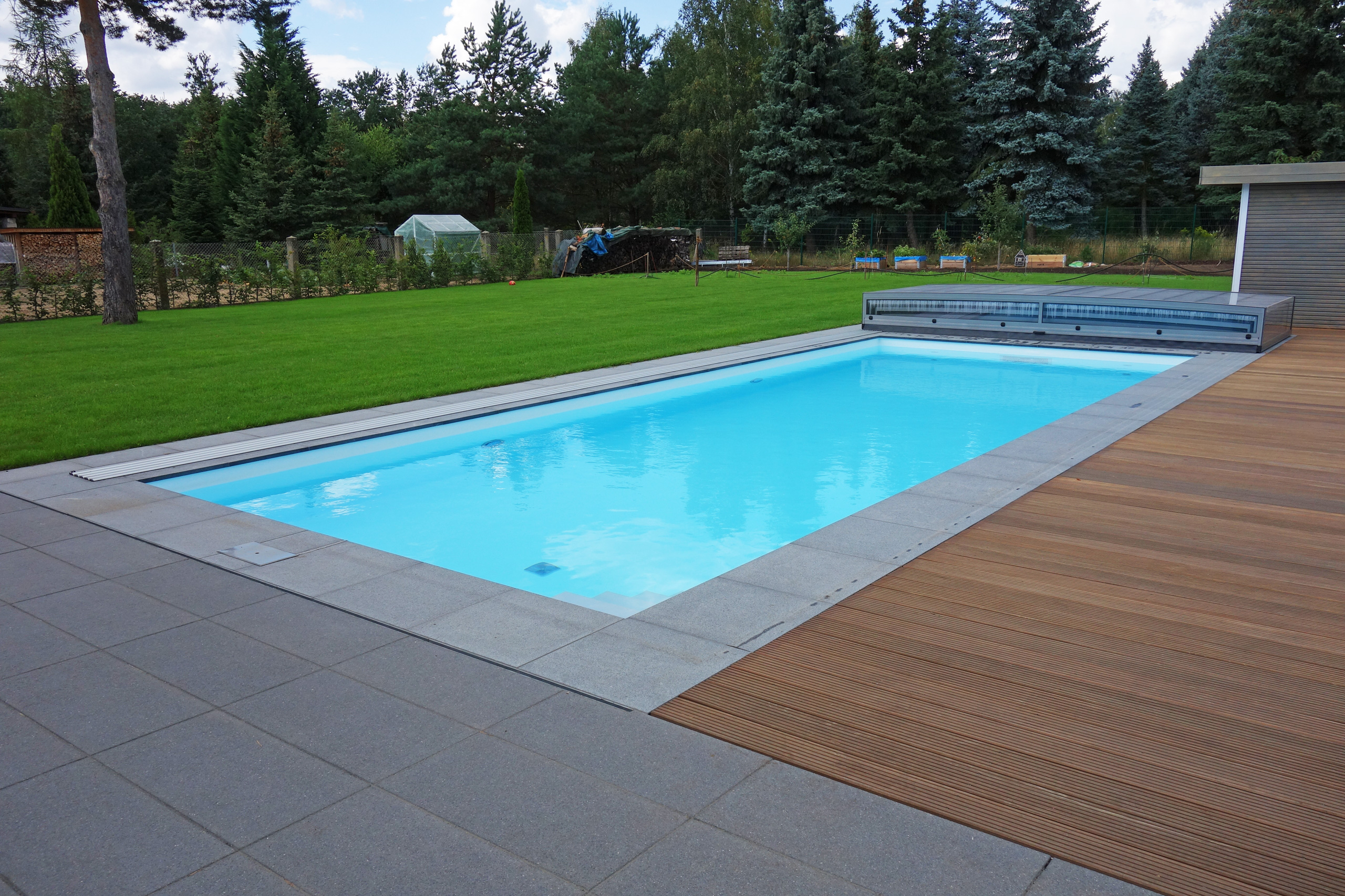 Detailansicht des Außenpools mit grauen Fliesen und Holzterrasse, umgeben von Rasen und Bäumen.