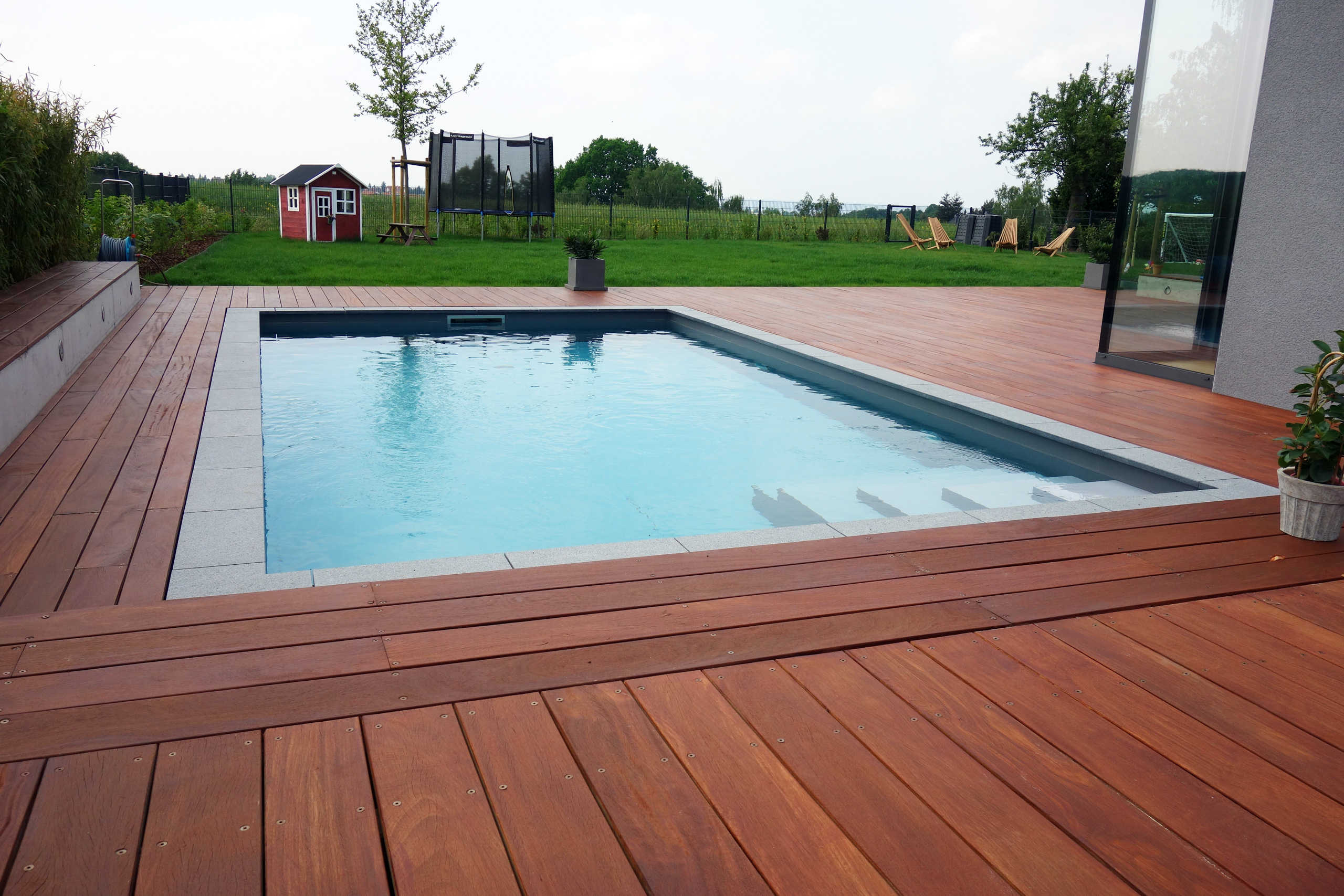 Holzdeck und Beckenrand aus Granitplatten um ein Betonbecken mit Ecktreppe und Podest