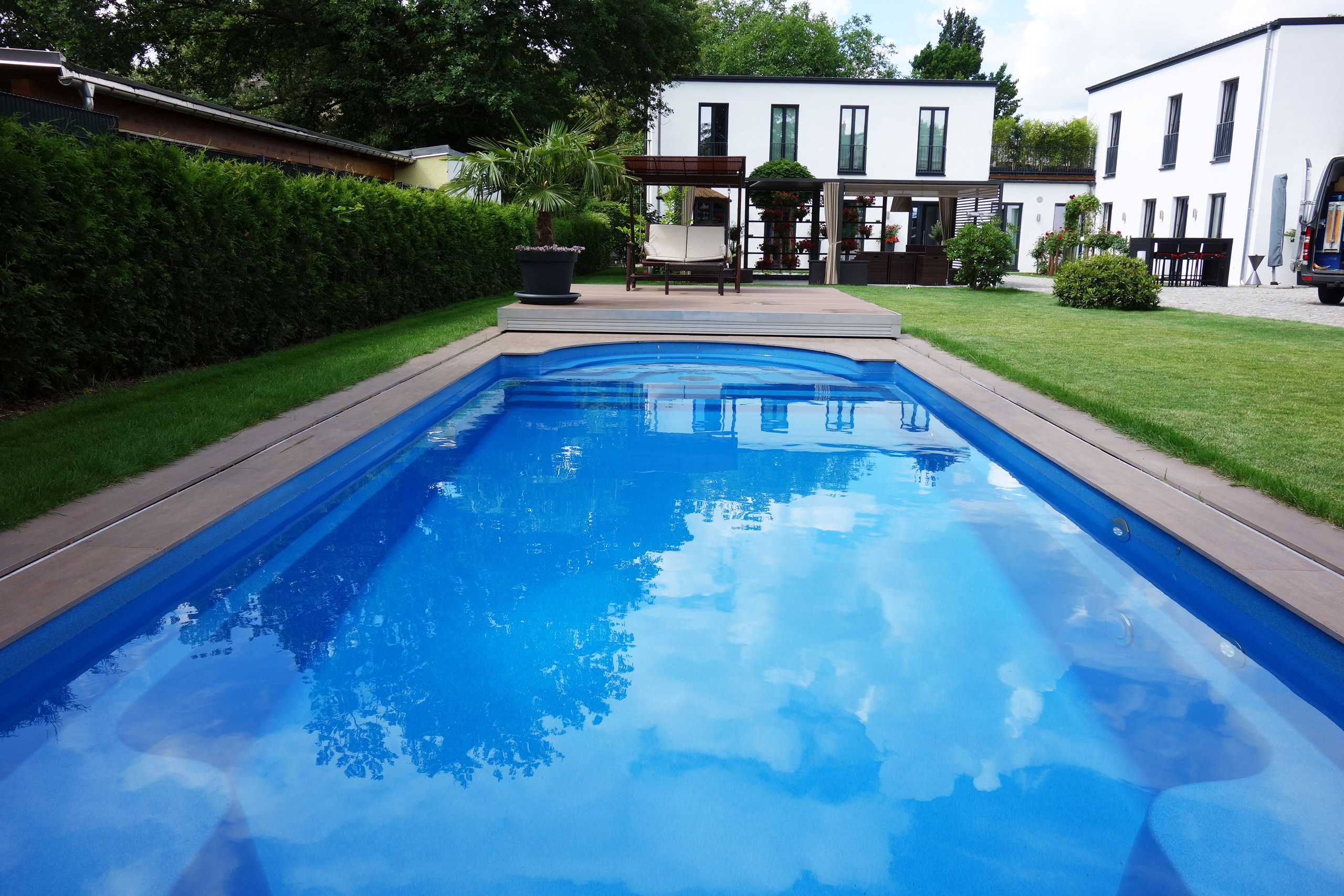 Blick auf das blaue Compass Pool Schwimmbecken Java 101 mit Terrassenabdeckung und Gartenanlage.