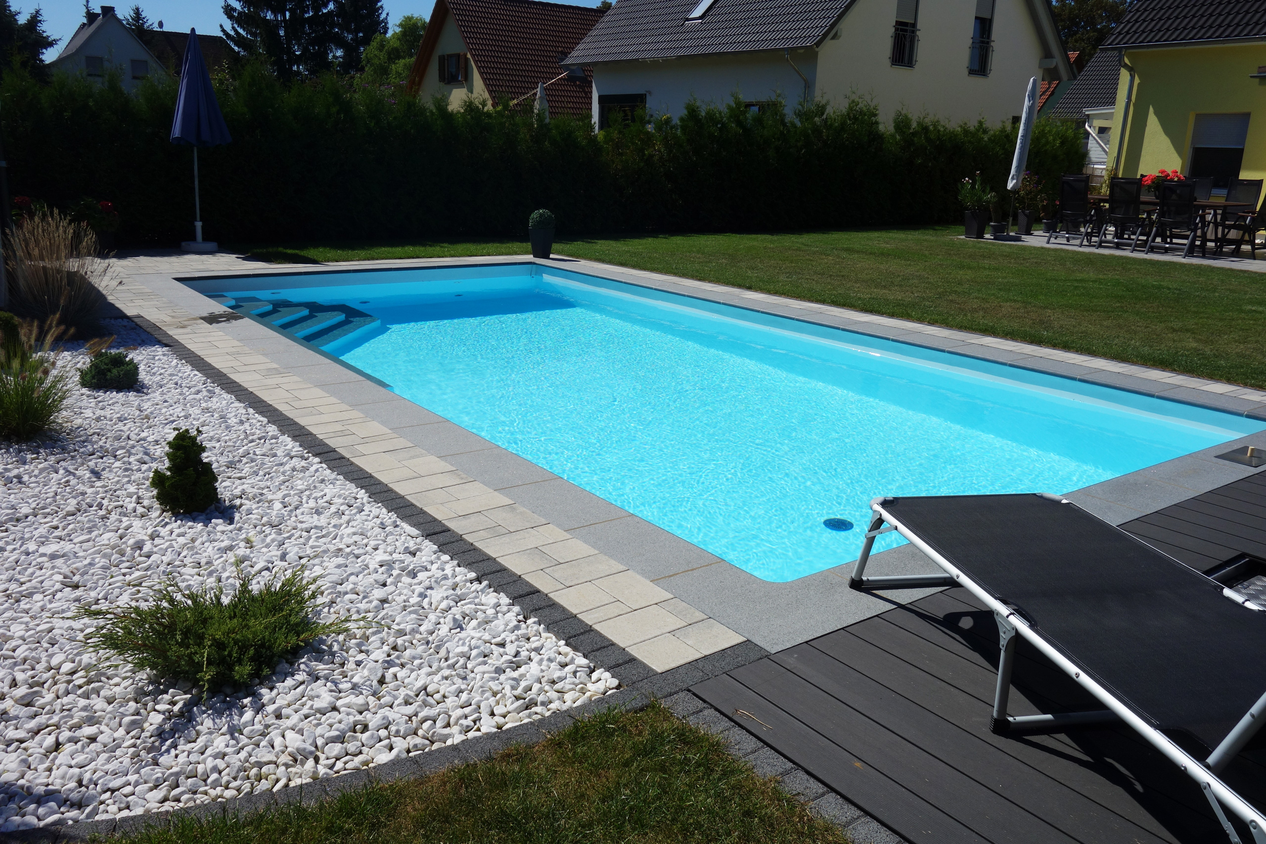 Rechteckbecken mit Ecktreppe, Podest und Luftsprudelsitz in einem Garten mit Liege und weißem Kies.