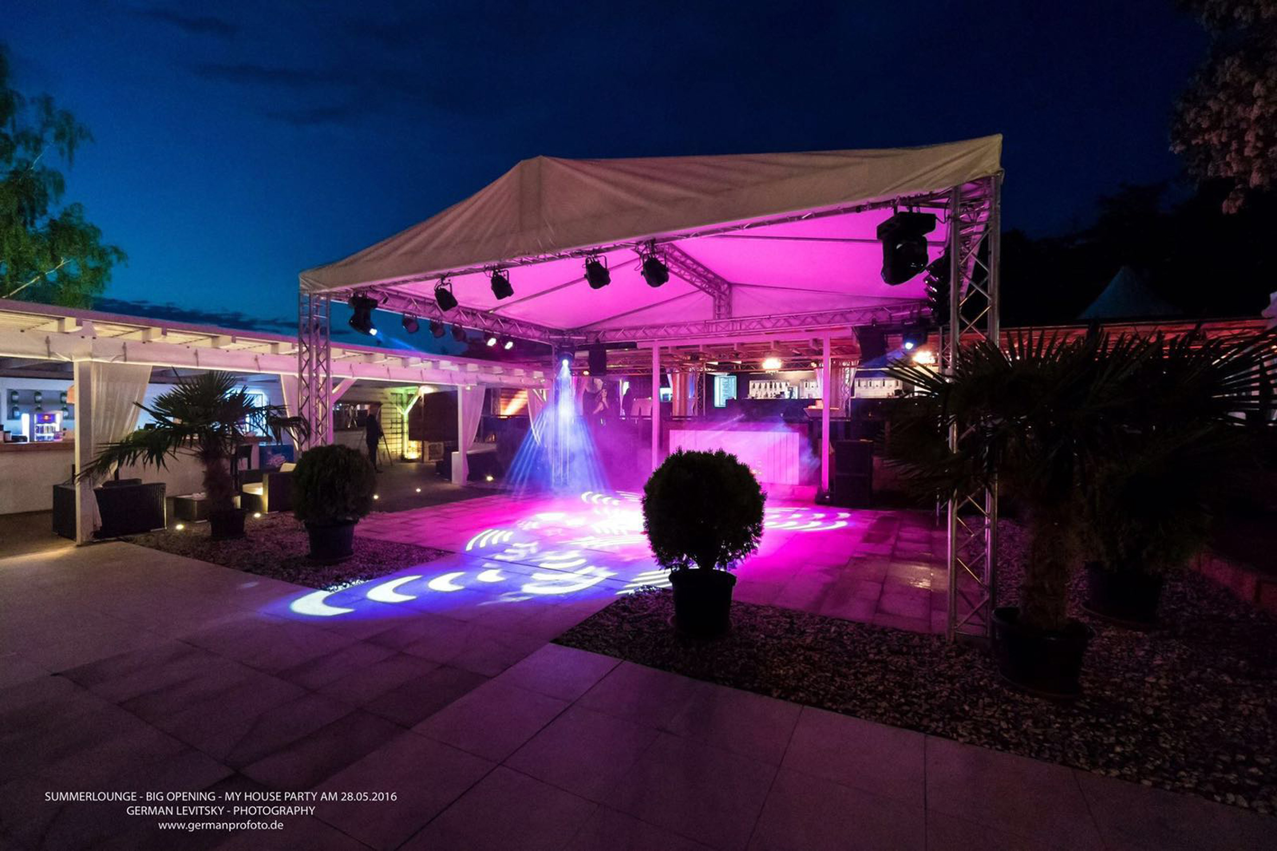 Beleuchteter Partybereich mit Zelt, DJ-Pult und Tanzfläche in Pink- und Blautönen bei Nacht